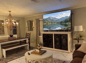 A living room with a flat-screen TV mounted above a fireplace, a dining area with a bench, and a coffee table with a remote control. - The Magnolia on Critz • Private Pool • Upscale Stay Near MSU (Starkville)