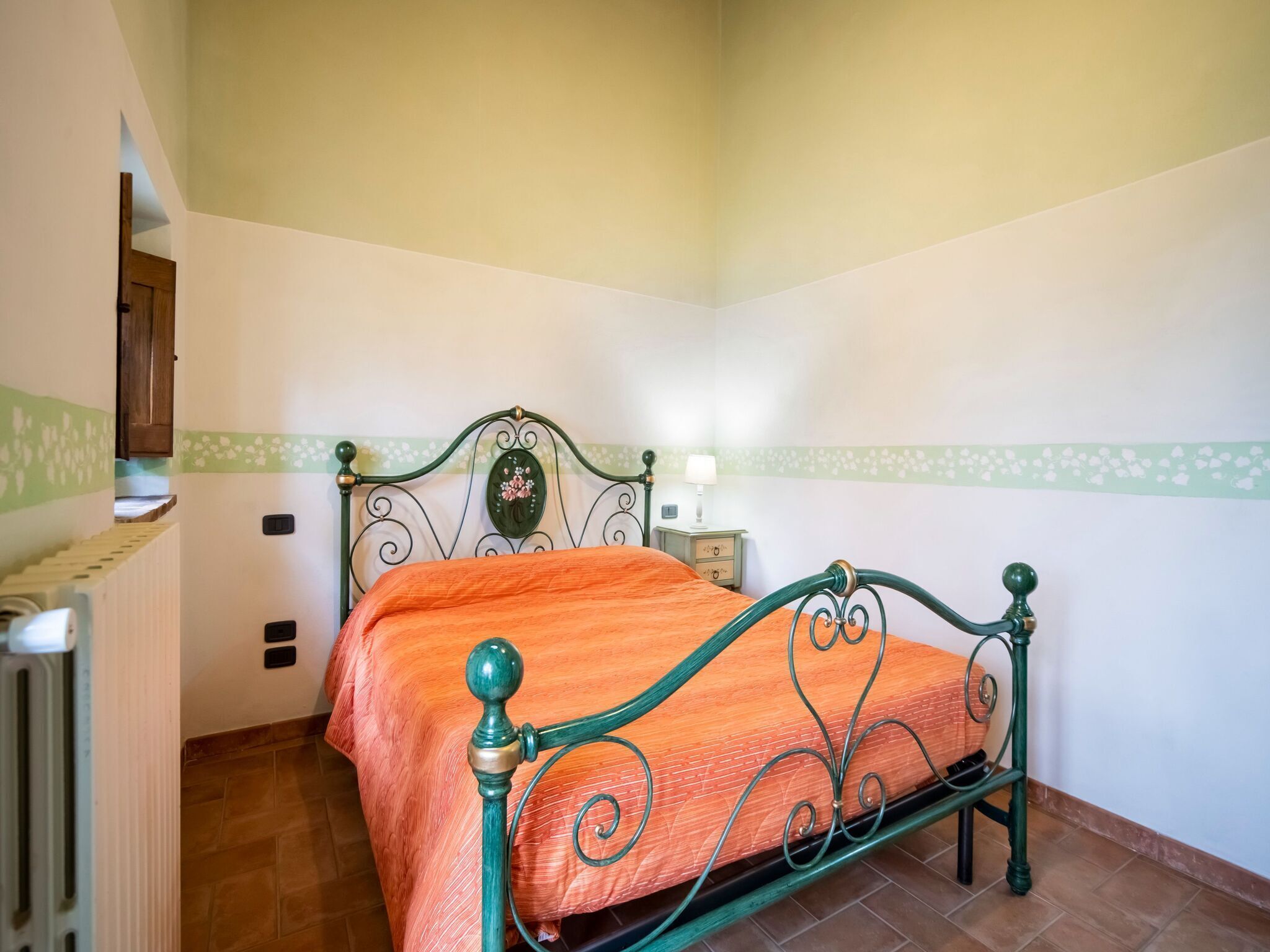 Una cama individual con estructura metálica verde y colcha naranja en una habitación de paredes blancas y borde floral verde.