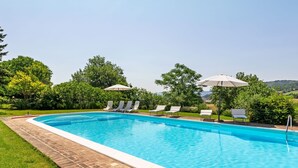 Una piscina rectangular con borde de ladrillo, rodeada de césped y árboles. Hay sillones de descanso y sombrillas a lo largo de la piscina.