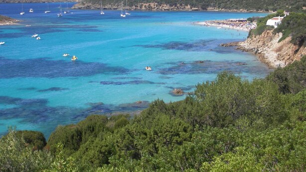 Una zona costera con aguas turquesas cristalinas, embarcaciones y una playa con gente.
