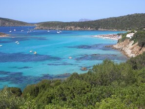 A coastal area with clear turquoise waters, boats, and a beach with people. - Belvilla by OYO Mura Pranu (Sant'Antioco)