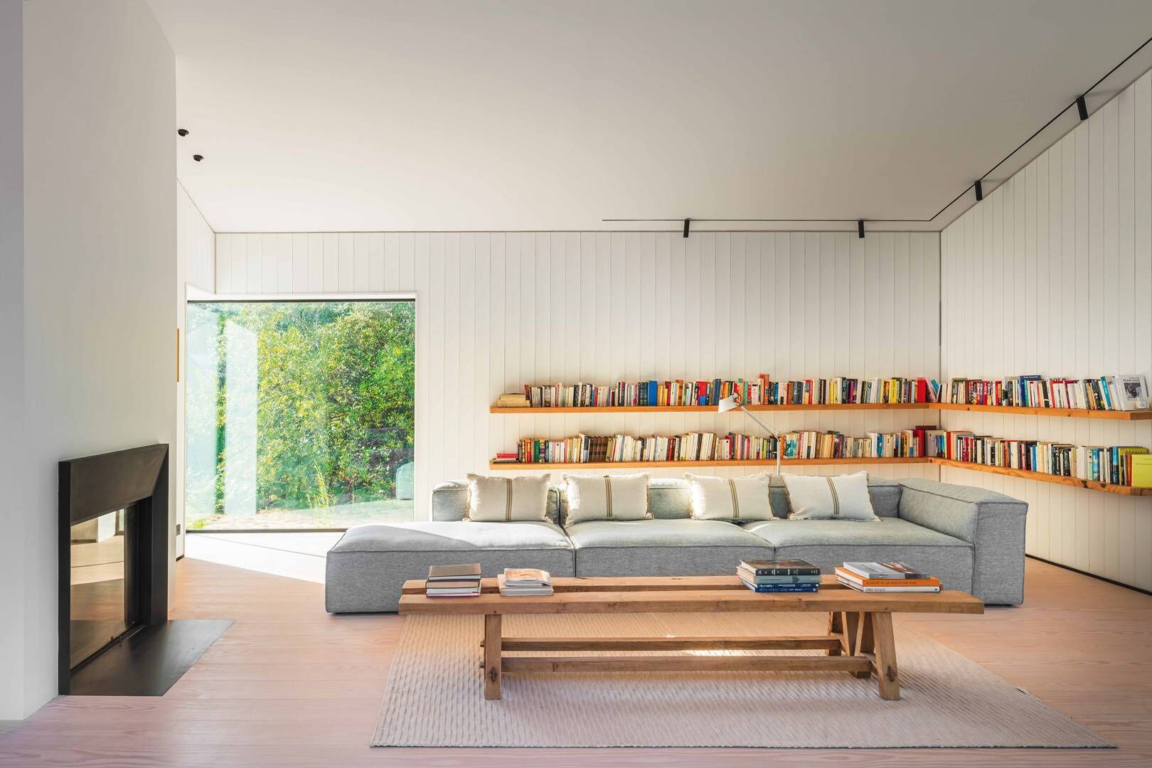 Een moderne woonkamer met een grijze hoekbank, houten boekenkasten, een houten salontafel en een groot raam met uitzicht op groen.