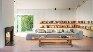 Un salon moderne avec un canapé d’angle gris, des étagères en bois, une table basse en bois et une grande fenêtre donnant sur un paysage verdoyant.