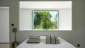 Une chambre avec une grande fenêtre, un lit avec des oreillers à rayures, une lampe sur une table de chevet et une porte au fond.