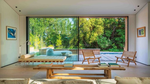 Un intérieur moderne avec une grande porte-fenêtre coulissante donnant sur une terrasse, un banc en bois et deux fauteuils en osier.