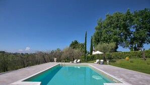 Ein rechteckiger Pool mit gewölbter Kante, umgeben von einer gefliesten Liegefläche. Im Hintergrund sind Liegestühle und ein weißer Sonnenschirm zu sehen.