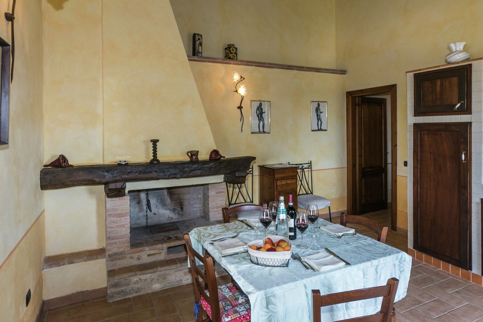 Un coin repas avec une cheminée en briques, une table dressée avec des verres à vin et un panier de fruits, et une porte en bois en arrière-plan.