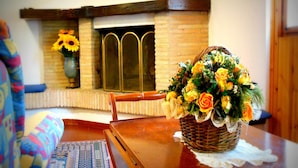 A room with a wooden table, a wicker basket with flowers, a chair, a sofa, a patterned rug, and a brick fireplace with a window.