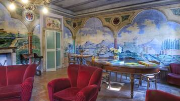 Una stanza con poltrone in velluto rosso, un tavolo di legno con libri, un lampadario e un affresco sulla parete.