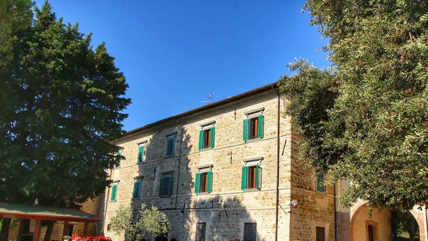 Un edificio de piedra con numerosas ventanas y contraventanas verdes, rodeado de árboles y mobiliario exterior.