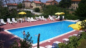 Una piscina con lettini e ombrelloni, circondata da edifici e vegetazione.