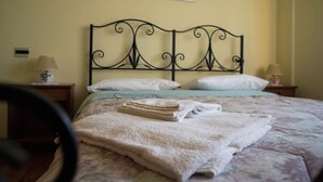 Un letto rifatto con cura, con una coperta a motivi floreali, un asciugamano bianco piegato e una testiera in metallo nero. Sono presenti due comodini con lampade e un quadro incorniciato appeso al muro sopra il letto.