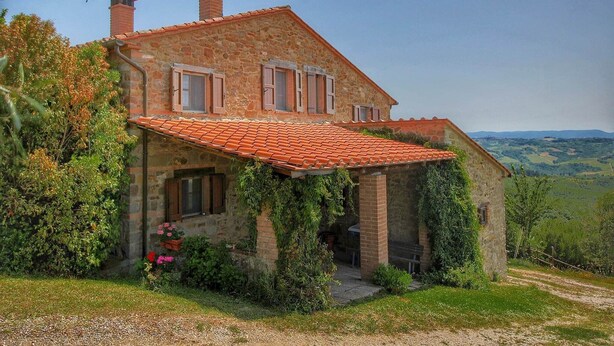 Une maison en pierre avec un toit recouvert de tuiles rouges, une cheminée et un jardin agrémenté de fleurs et de verdure.