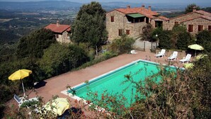 Un espace piscine agrémenté de chaises longues et de parasols, entouré de bâtiments en pierre aux toits de tuiles rouges, le tout se dressant sur un fond de montagnes.