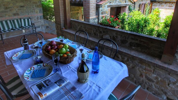Un espacio de comedor al aire libre con una mesa dispuesta para dos personas, con una cesta de frutas, copas de vino y una botella de agua.