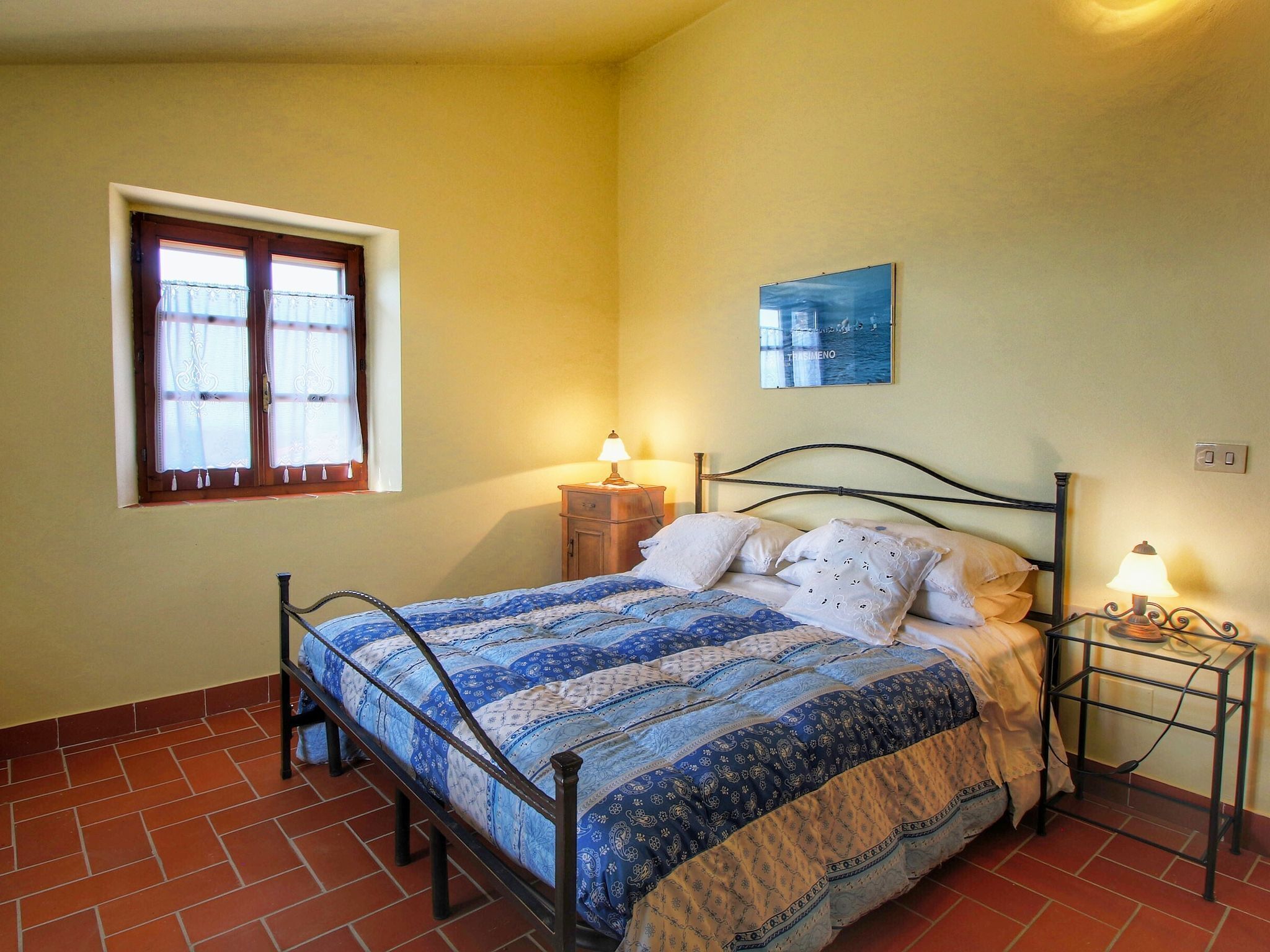 A bedroom with a bed, two bedside tables, a window with curtains, and a wall-mounted picture.