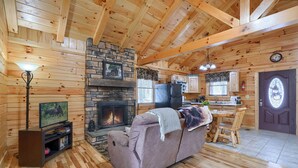 A cozy cabin interior with a stone fireplace, wooden walls, a sofa, a television, and a dining area.