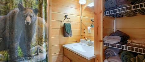 Une salle de bain rustique, aménagée dans une cabane en bois, avec une fresque représentant un ours, un lavabo et des étagères où sont rangées des serviettes pliées.