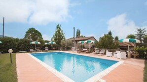A rectangular swimming pool with tiled edges, surrounded by lounge chairs and umbrellas, set against a backdrop of trees and a stone building. - Cottage in Magione near Lake Trasimeno (Magione)