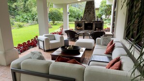 Un espace extérieur aménagé, abrité et doté d’une cheminée en pierre, de meubles en osier et offrant une vue sur un jardin luxuriant.