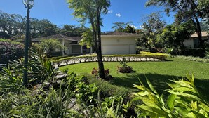 A house with a garage, surrounded by a well-maintained garden with various plants and a clear blue sky. - Boquete Luxury: Chic & Serene Home, Walk to Town & Amenities (Boquete)