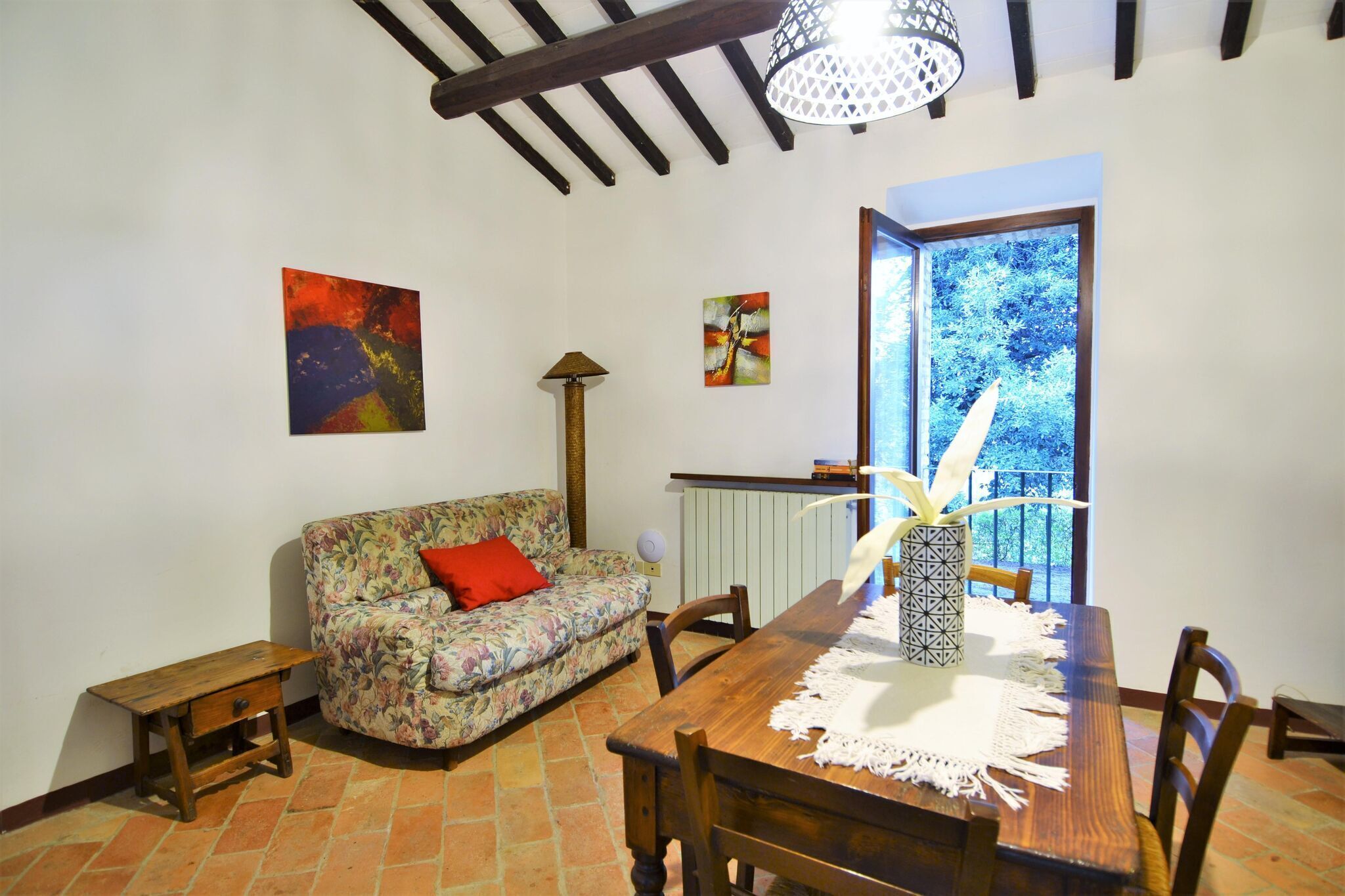 A dining area with a wooden table, chairs, a floral sofa, and a window with a view of trees.