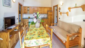 Ein gemütliches Zimmer mit Holzmöbeln, einem Esstisch mit blumigem Tischläufer, einer Couch und einem Fernseher auf einem Holzständer.