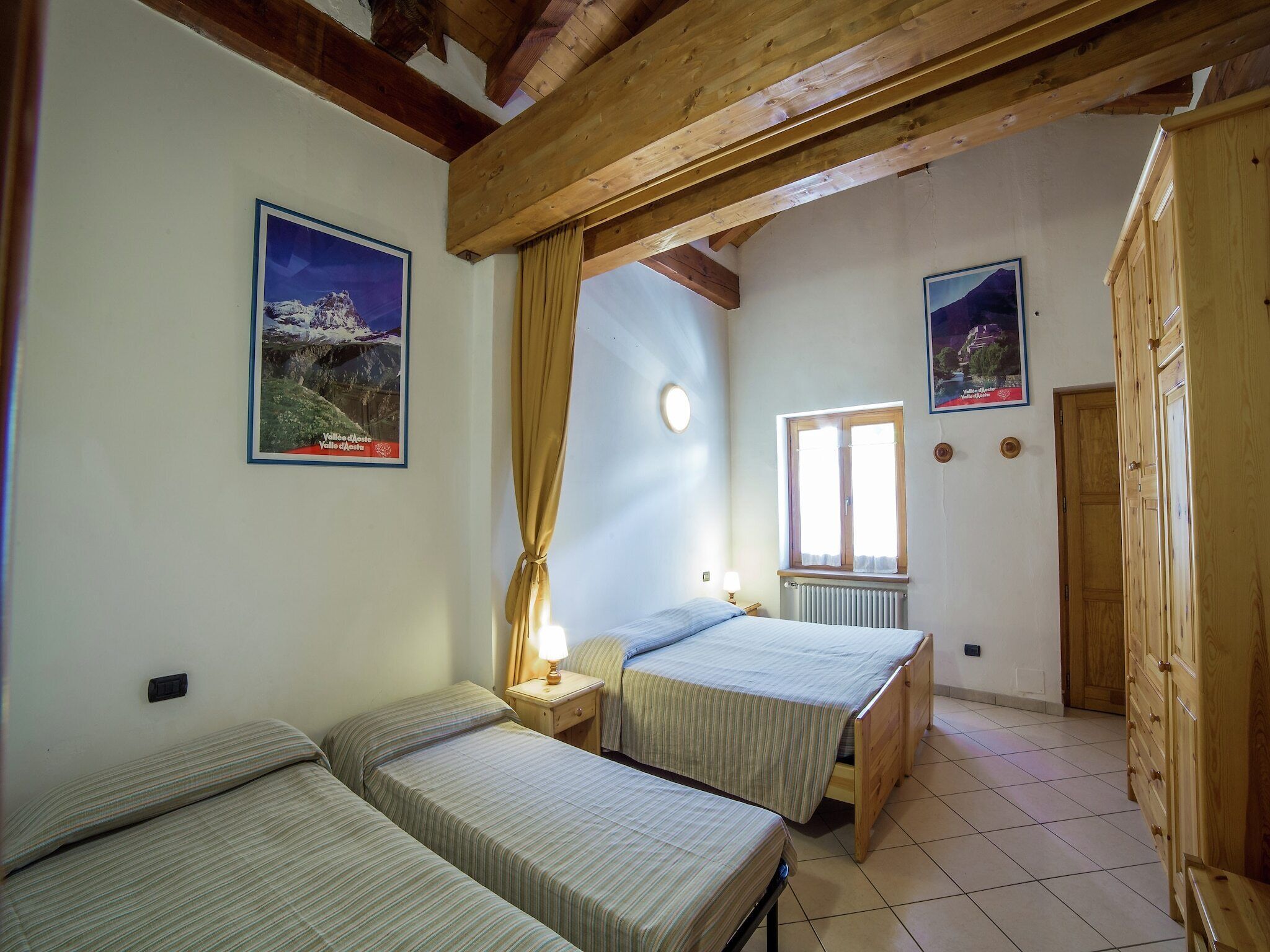 Une chambre avec deux lits simples, des poutres en bois au plafond, une fenêtre avec des rideaux et deux tableaux encadrés accrochés aux murs.