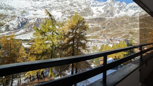 A mountain view from a balcony with a railing, snow-covered peaks, and a town below.