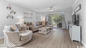 A modern living room with a sofa, coffee table, and TV stand. There are shelves with decorative items and a large window with a view.