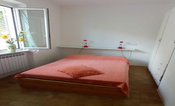 Une chambre avec un lit simple, une fenêtre avec des rideaux, un radiateur et une étagère sur laquelle reposent deux objets rouges.