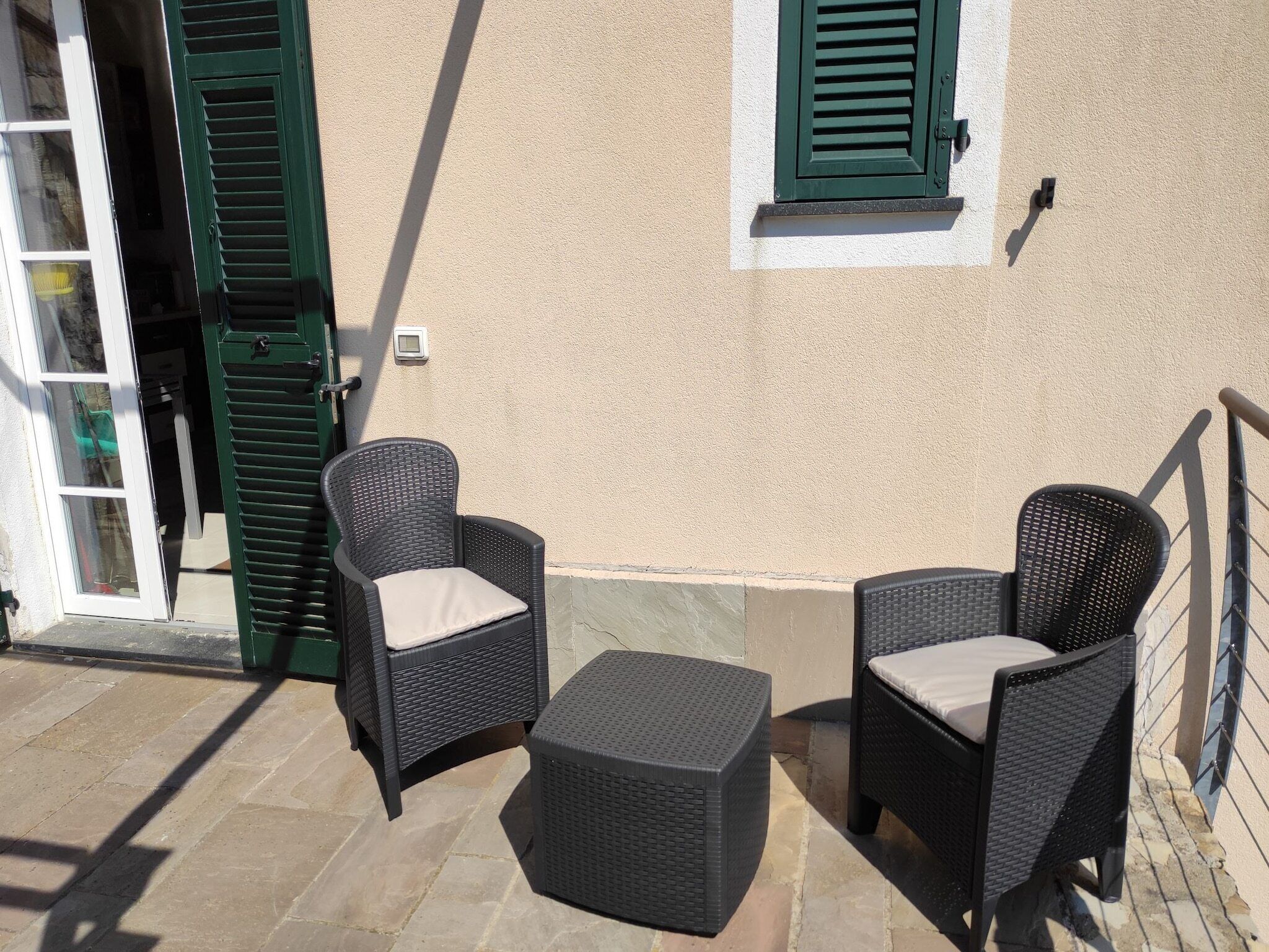 An outdoor seating area with wicker furniture and a small table, adjacent to a building with green shutters and a beige wall.