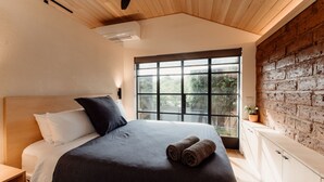 A bedroom with a large bed, a wooden ceiling, a brick wall, and a window with a view of greenery. - Great 50 meter square architectural fitout with a cosy modern vibe to relax in  (Surf Beach)