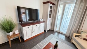 A living room with a flat-screen TV, a cabinet, a plant, a small table, a sofa, and a window with curtains.