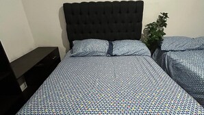Una cama con colcha estampada, cabecera oscura y una planta en la mesita de noche.