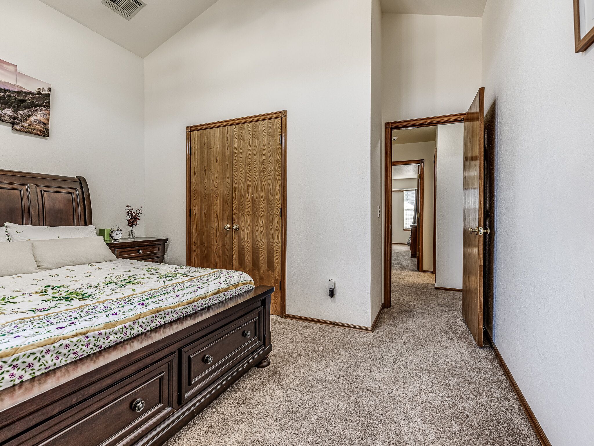 A bedroom with a large bed, a wooden wardrobe, and a hallway leading to another room.