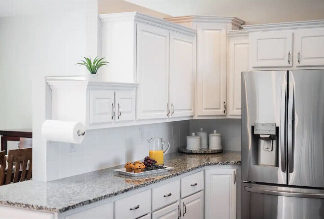 A kitchen with white cabinets, a stainless steel refrigerator, a granite countertop, a plant, and a fruit bowl.