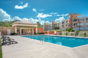 Ein Schwimmbecken mit Liegestühlen und einer überdachten Sitzgelegenheit, umgeben von mehrstöckigen Gebäuden mit Balkonen.