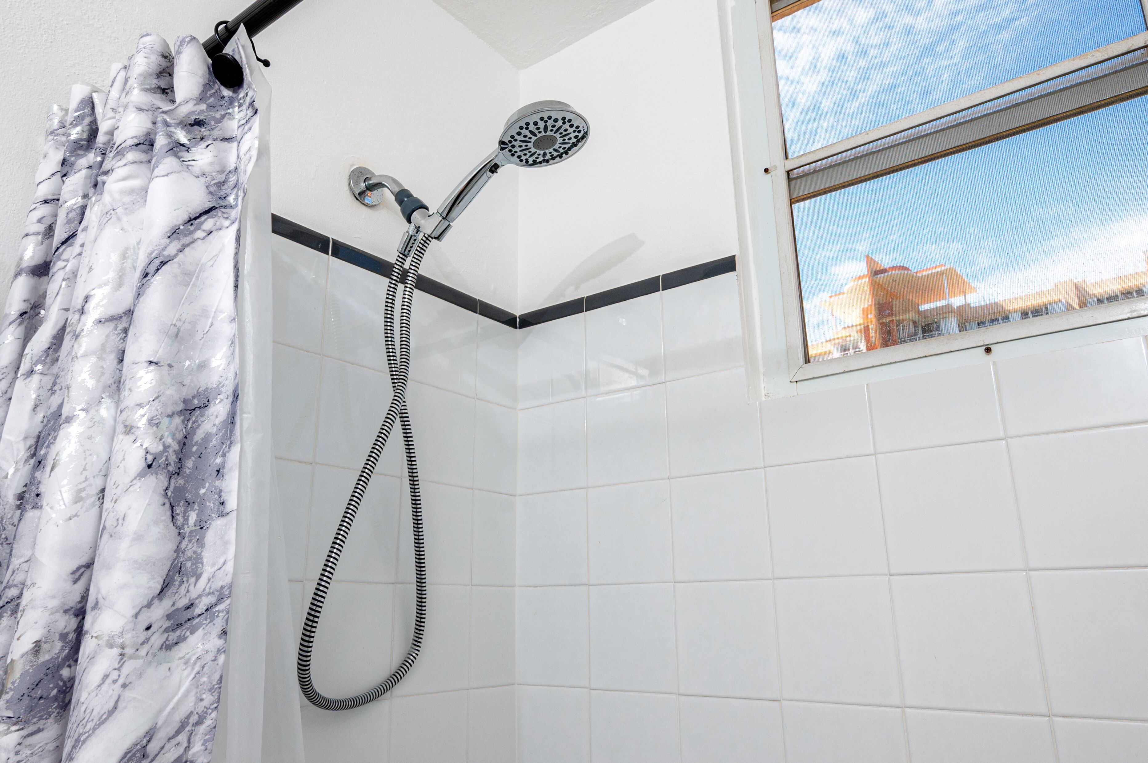 A shower with a handheld showerhead and a flexible hose, a marble-patterned shower curtain, and a window with a view of a building.