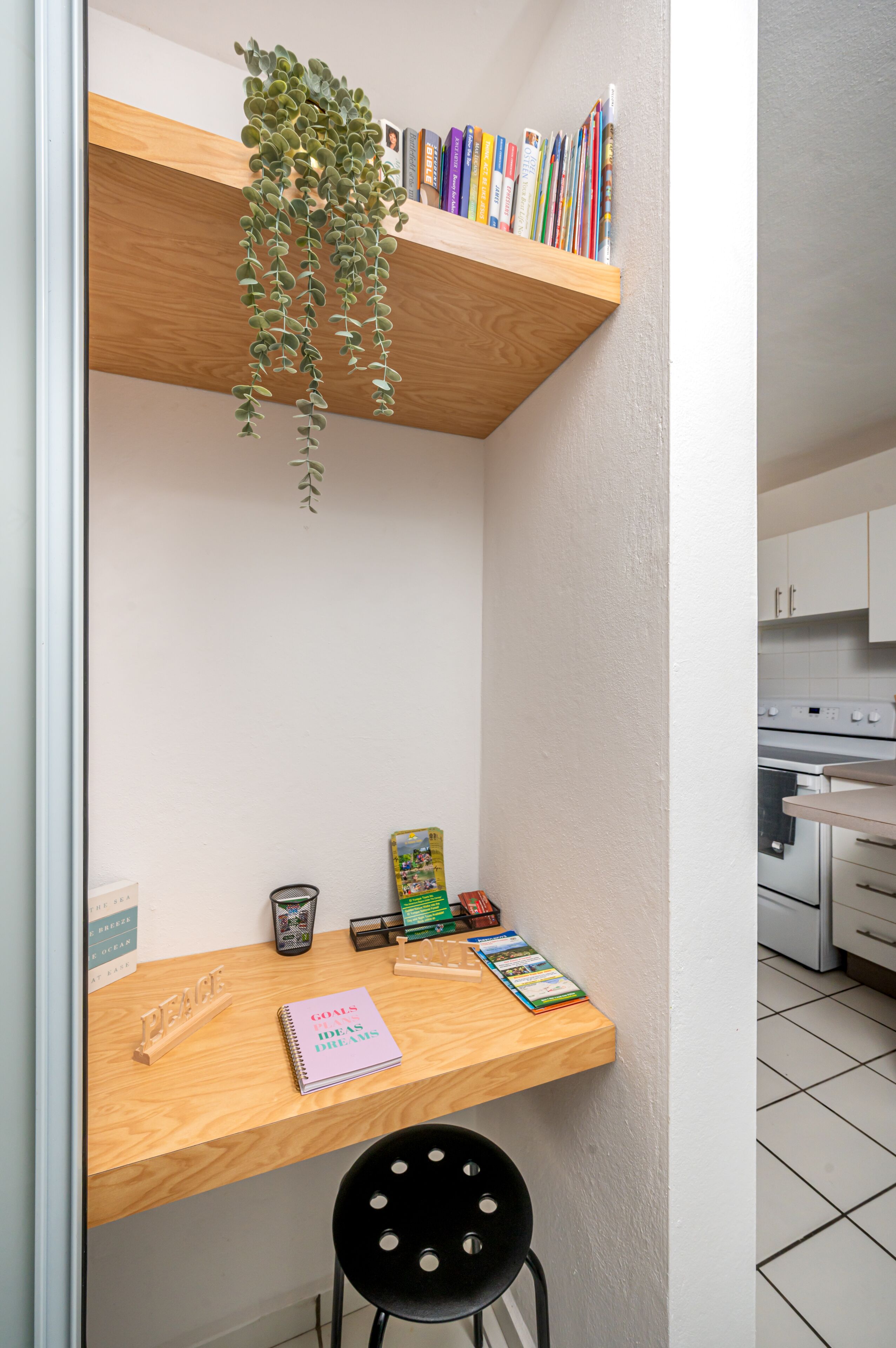 A compact room with a wooden desk, a black chair, a bookshelf with books, a hanging plant, and a small trash bin.