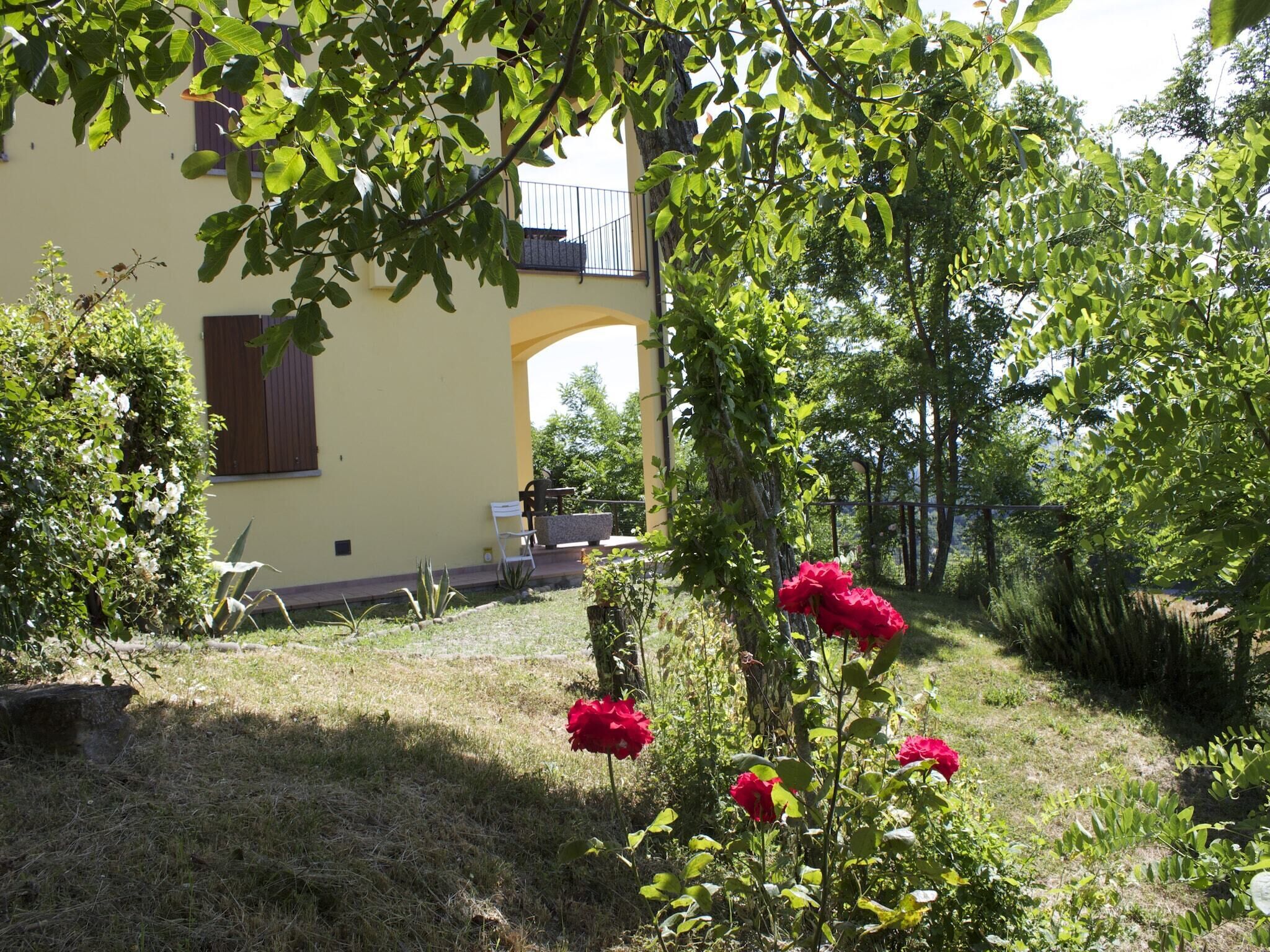 A garden with a yellow building, a wooden bench, and a red rose bush.