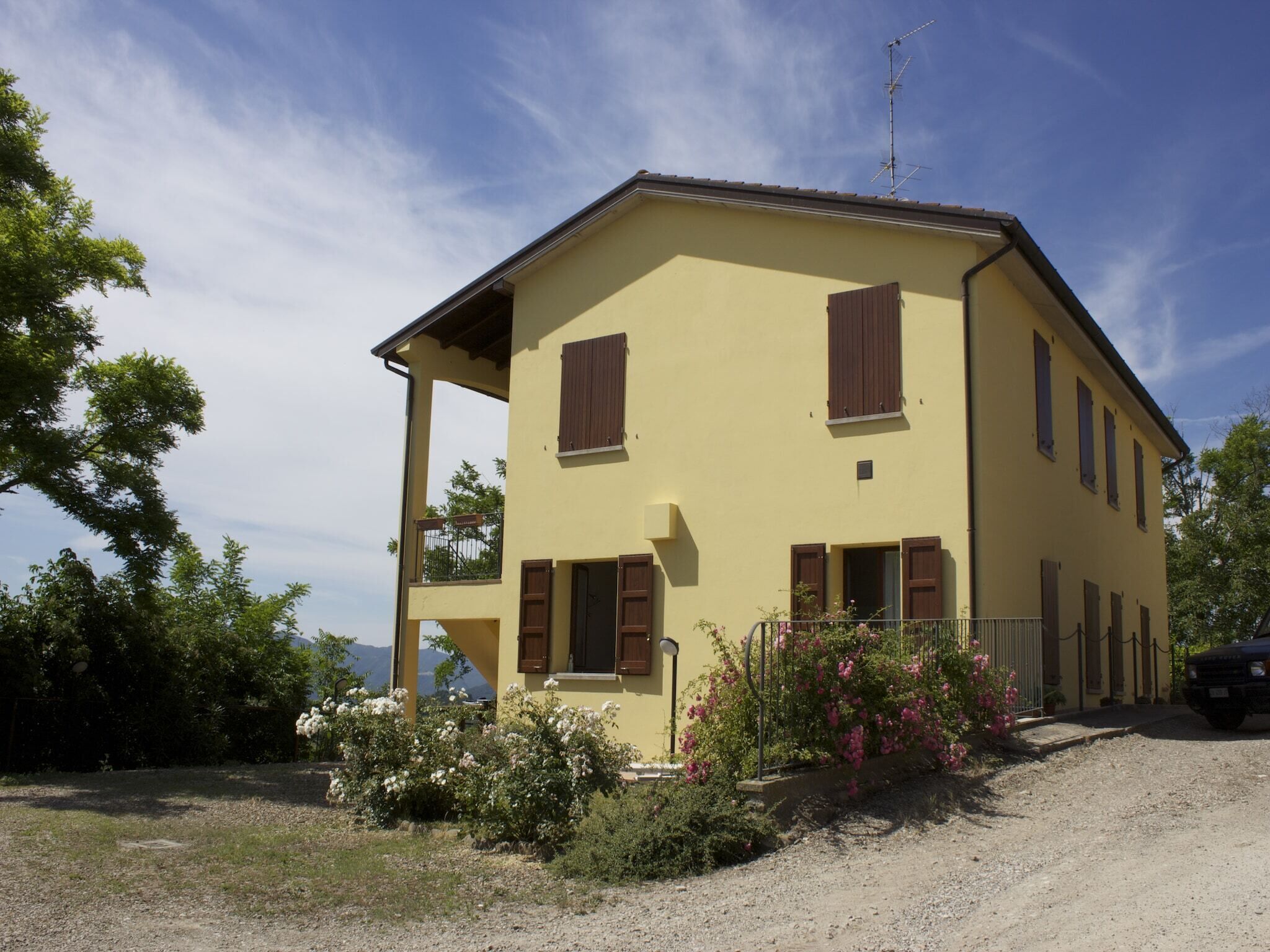 A two-story yellow building with brown shutters, a balcony, and a gravel driveway.