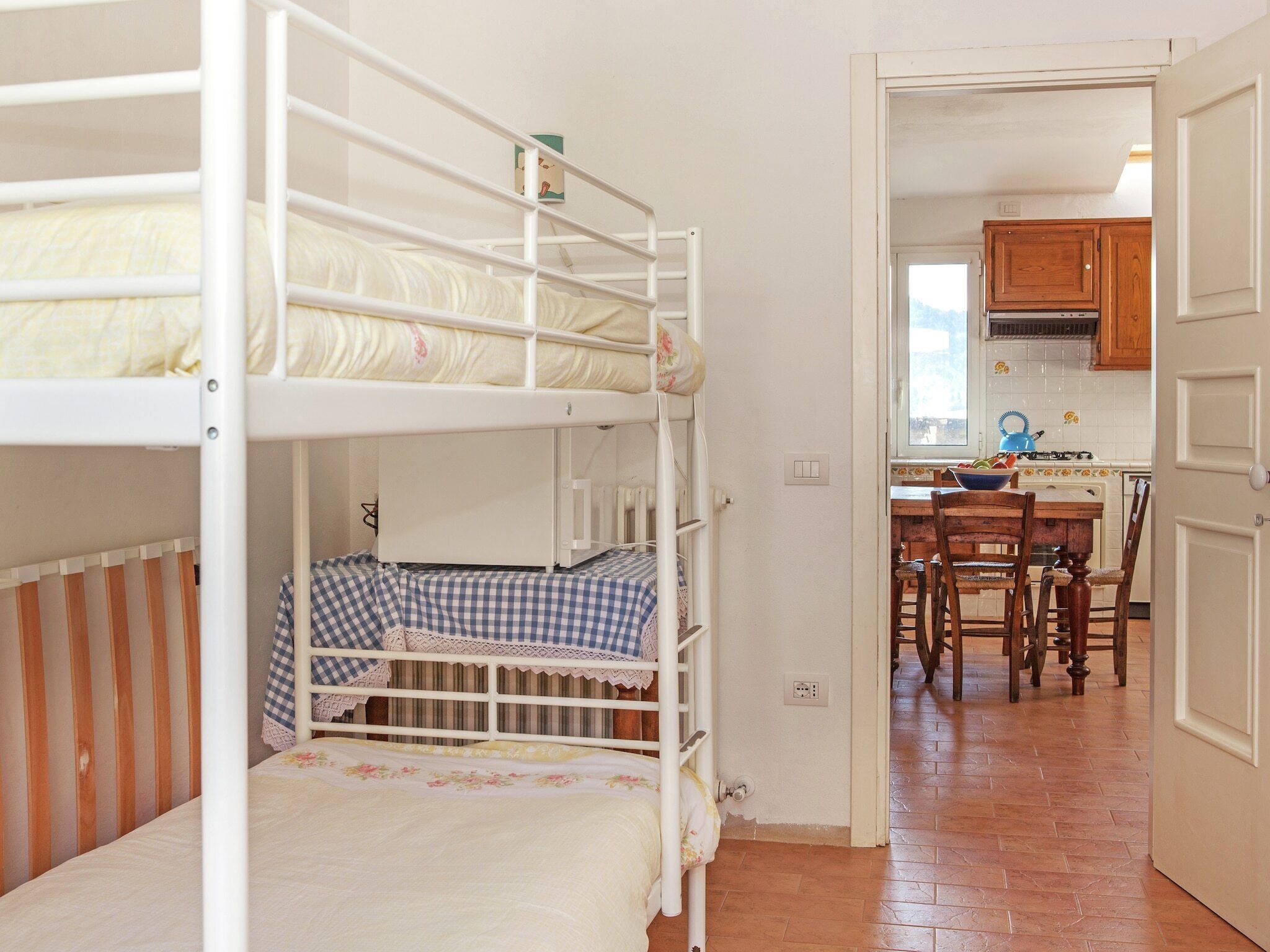 A bunk bed room with a checkered bedspread, a wooden crib, and a kitchen area with wooden cabinets and a tiled backsplash.