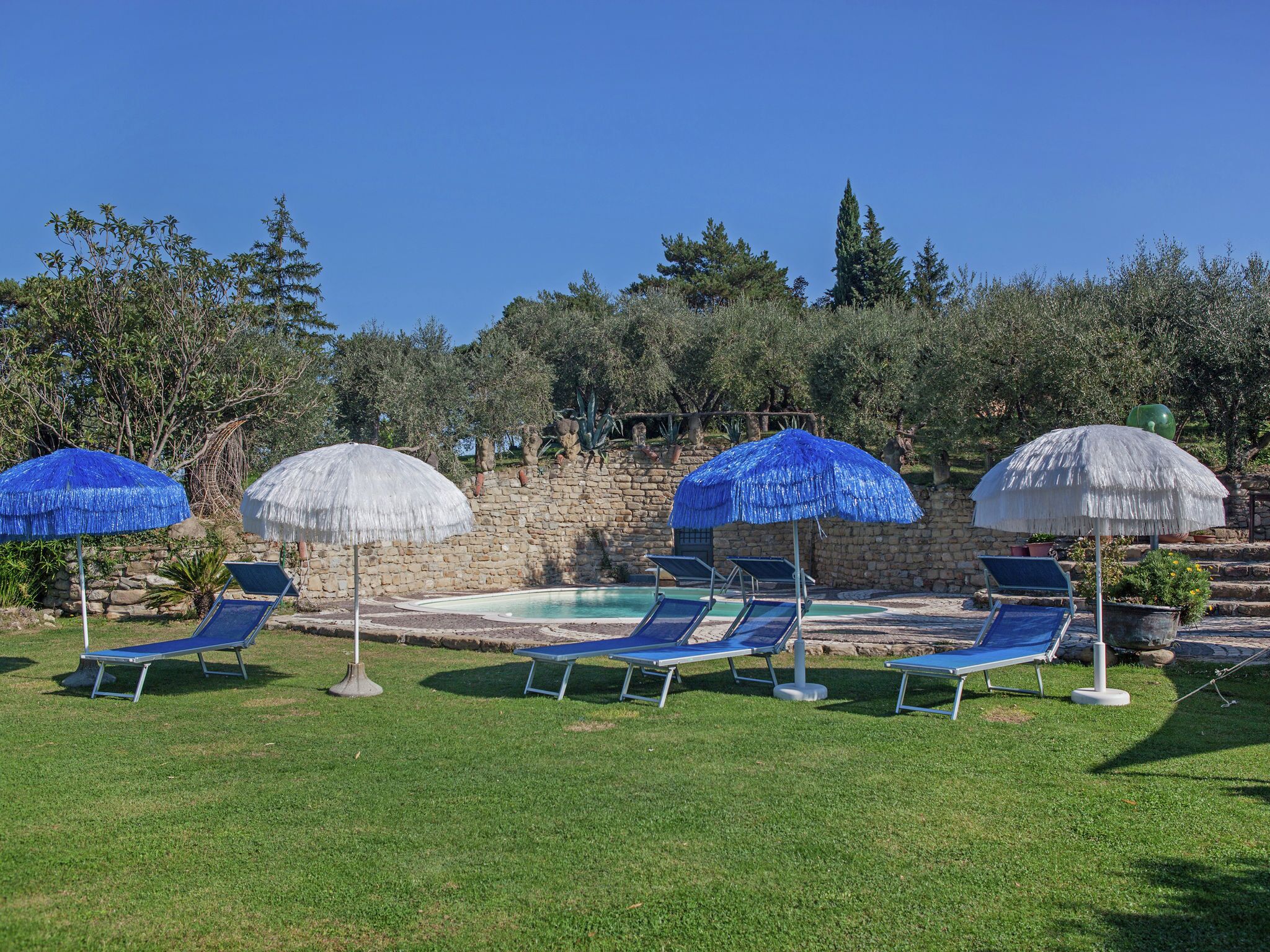 A poolside area with lounge chairs and umbrellas, surrounded by stone walls and greenery.