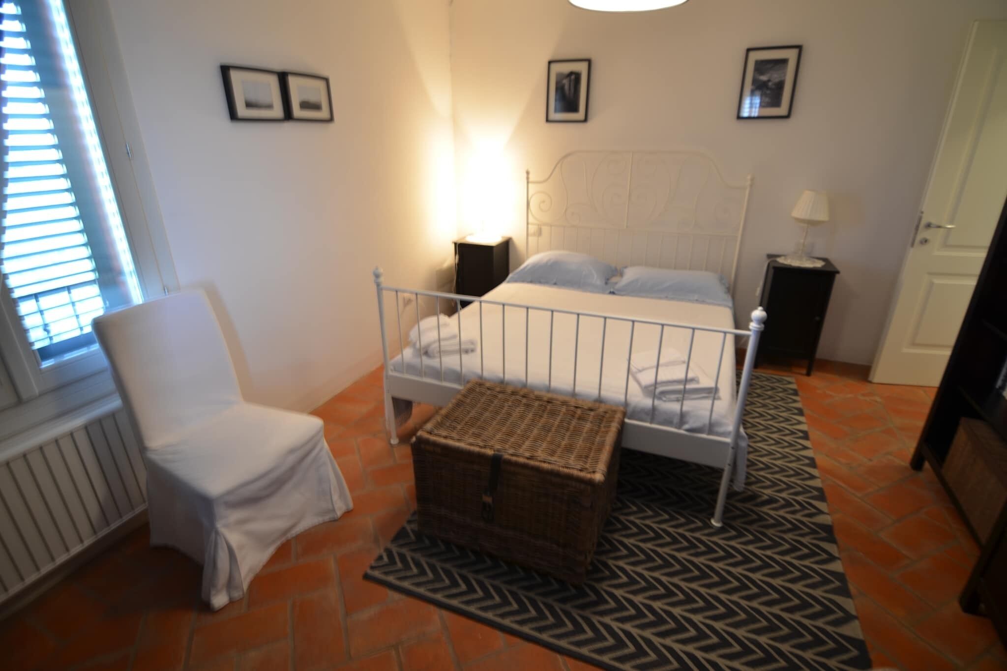 Un dormitorio con una cama de metal blanca, una silla blanca, una cesta de mimbre y una alfombra estampada.