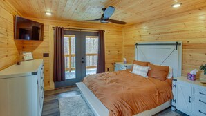 Un dormitorio de cabaña de madera con cama, ventilador de techo, televisión y puerta corrediza al exterior.