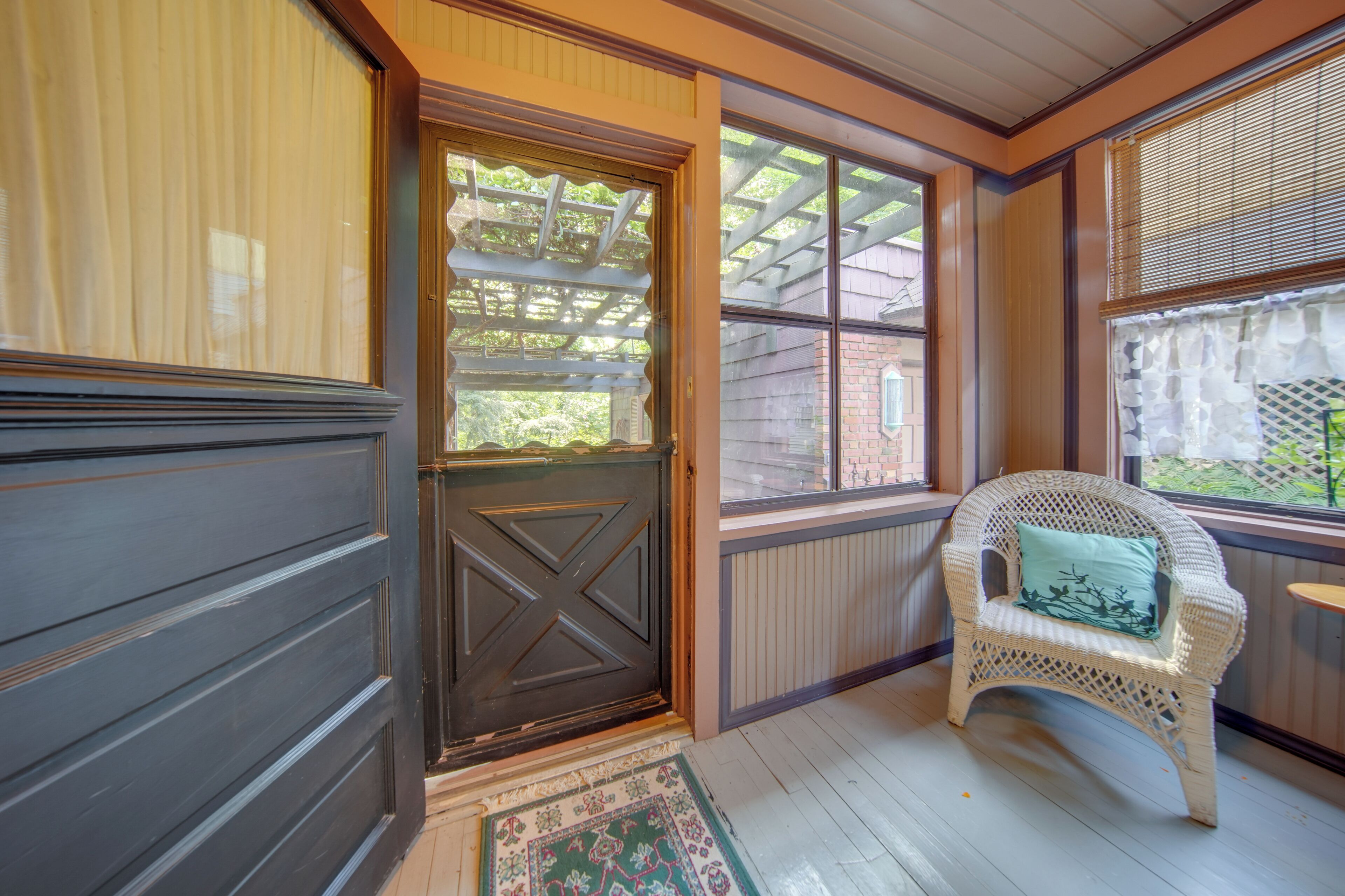 A porch with a dark wooden door, a wicker chair with a cushion, and a view of a brick building through the windows.