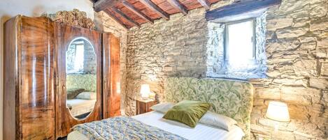 Une chambre à coucher au style rustique, avec un mur en pierre, des poutres apparentes en bois au plafond, un lit recouvert d’une literie à motifs floraux, une armoire en bois et une petite fenêtre.