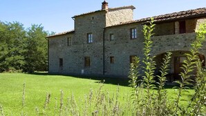 Un edificio in pietra con tetto in tegole, un prato e una piccola area giardino con piante alte.