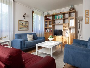 A living room with a blue sofa, a white coffee table, and a TV. There is a bookshelf with decorative items and a painting on the wall.
